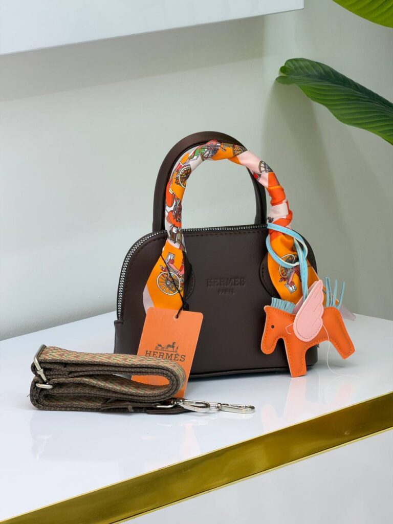 کیف مینی هرمس کد ۹۳۷ قهوه‌ای با دسته پیچیده شده در اسکارف نارنجی و آویز اسب پگاسوس روی میز سفید